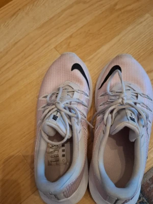 Nike vita och rosa sneakers - Snygga sneakers från Nike i vitt och ljusrosa med svart swoosh på ovansidan. Skorna har meshmaterial för bra ventilation, vita snören och en slitstark sula med grepp. Perfekta för dig som gillar sportig stil och komfort.