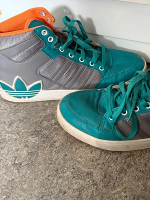 Vintage Adidas high top sneakers - Vintage Adidas high top sneakers i teal, grått och vitt med orange insida. Klassiska tre ränder på sidan och stor Adidas-logga bak. Skorna har snörning, vit sula och är gjorda i en mix av textil och syntetmaterial. Köpta från beyond retro, storlek 40