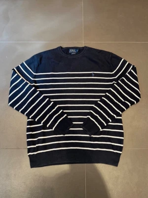 Marinblå randig tröja Polo Ralph Lauren - Snygg marinblå stickad tröja från Polo Ralph Lauren med vita horisontella ränder och klassisk broderad logga på bröstet. Rund halsringning och ribbade muddar. Perfekt till jeans eller chinos för en clean look.