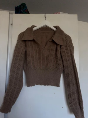 Brun stickad tröja med krage - Säljer en brun stickad tröja med bred krage och v-ringning. Tröjan har ett ribbat mönster, lång ärm och en croppad passform med vågig nederkant. Perfekt för dig som gillar en trendig och mysig look.