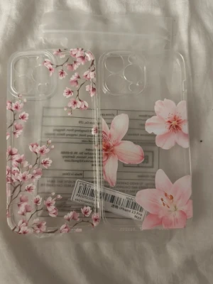 Genomskinliga mobilskal med blommor 13 pro max - Två genomskinliga mobilskal med rosa blommönster. Ena skalet har små körsbärsblommor över hela ytan, medan det andra har större blommor i rosa nyanser. Skalens design är tunn och flexibel, perfekt för dig som vill ha ett snyggt och stilrent skydd till mobilen.