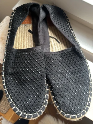 Svarta espadrilles med flätad ovandel - Säljer ett par svarta espadrilles med flätad textilovandel och små vita prickar. Skorna har en klassisk sula i rep och dekorativa sömmar runt kanten. Perfekta för sommaren och riktigt sköna att bära. Passar 38 med