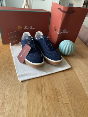 Marinblå sneakers från Loro Piana - Snygga marinblå sneakers från Loro Piana med ovandel i mocka, vita detaljer vid hälen och en klassisk gummisula. Skorna har matchande marinblå skosnören och en stilren, låg profil som passar till många olika outfits.