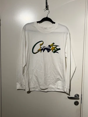 Vit långärmad t-shirt med tryck - Vit långärmad t-shirt med färgglatt tryck på bröstet i grönt, gult och blått. Trycket har en graffiti-liknande stil och inkluderar en stjärna och textdetaljer. Perfekt för en avslappnad och trendig look.