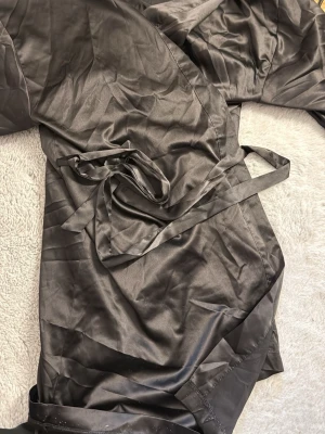 Svart satin morgonrock med skärp - Snygg svart morgonrock i glansigt satinmaterial med tillhörande skärp i midjan. Perfekt för att chilla hemma i och ger en lyxig känsla. Lång ärm och klassisk design.