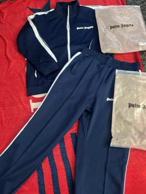 Mörkblå tracksuit från Palm Angels - Snygg mörkblå tracksuit från Palm Angels med vita ränder längs ärmar och ben. Setet består av zip-jacka med hög krage och matchande byxor med resår i midjan. Palm Angels-logga broderad på bröstet och benet. Perfekt för chill dagar hemma eller streetwear. Pris kan diskuteras och mer bilder kan skickas 