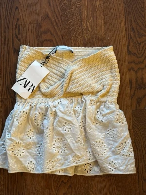 Beige och vit tubtopp från Zara - Trendig tubtopp från Zara i beige och vitt. Överdelen är ribbad och stickad medan underdelen har volang i broderad bomull med spetsdetaljer. Perfekt för sommarens alla looks och ger en romantisk vibe.
