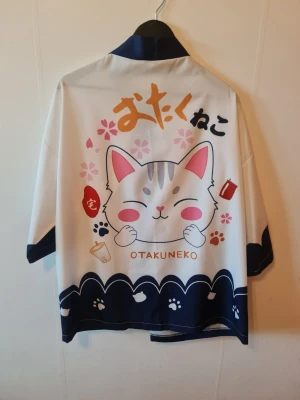 Kimono med kattmotiv Otakuneko - Japansk inspirerad kimono i vitt och marinblått med sött kattmotiv, körsbärsblommor och texten 'OTAKUNEKO'. Perfekt för cosplay eller anime-event. Materialet känns lätt och mjukt, har vida ärmar och kontrastfärgade detaljer.