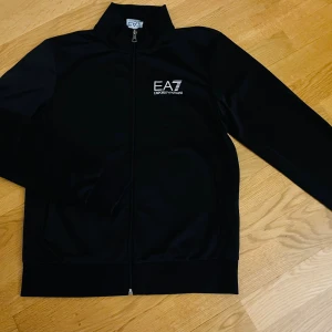 Svart track jacket från EA7 Armani - Svart track jacket från EA7 Emporio Armani med vit logga på bröstet och dragkedja hela vägen. Jackan har hög krage. Ny skick . 