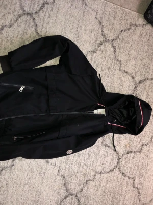 Svart vindjacka från Moncler S - Snygg svart vindjacka från Moncler med huva och dragkedja. Jackan har vita och röda detaljer längs huvan och insidan, samt två snedställda fickor med dragkedja. Tillverkad i ett lätt och slitstarkt material, perfekt för blåsiga dagar.