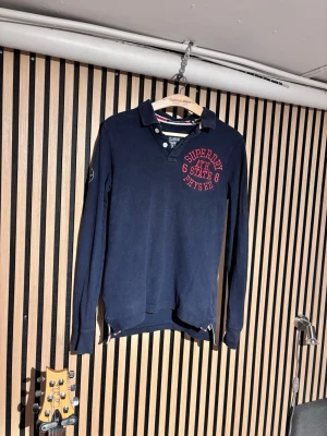 Mörkblå långärmad piké Superdry - Mörkblå långärmad pikétröja från Superdry med röd broderad text på bröstet. Klassisk krage med två knappar och diskreta detaljer vid sidorna. Perfekt för en avslappnad men snygg stil. Har ett litet hål i armen som man ser på bilden!