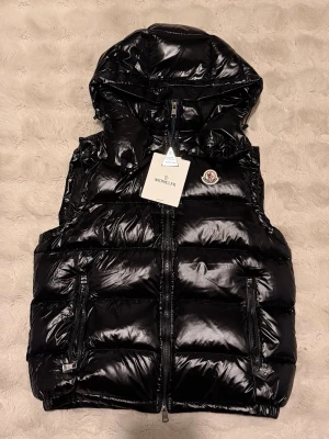 Svart dunväst från Moncler med huva - Snygg svart dunväst från Moncler med glansig finish och avtagbar huva. Västen har två dragkedjeförsedda fickor, klassisk Moncler-logga på bröstet och quiltad design. Perfekt för lager-på-lager och streetstyle.