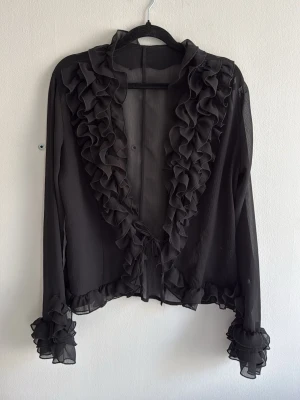 Svart transparent volangblus - Elegant svart blus i transparent material med volangdetaljer längs krage, framkant och ärmslut. Långa ärmar och knytband framtill. Perfekt för dig som gillar romantisk och trendig stil.