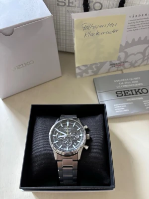 Seiko silverfärgad kronografklocka 100m - Stilren Seiko kronografklocka i silverfärgat rostfritt stål med svart urtavla och ljusblå detaljer. Klockan har tre subdials, datumvisning och robust länkarmband. Perfekt accessoar för dig som gillar klassisk men sportig stil. Köp i Kalmar. Allt på bilderna skickas med + extra länkar.