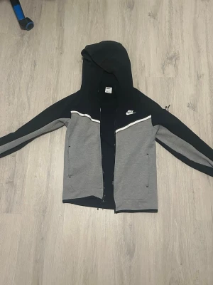 Niketech dress svart och grå - Hoodien för 699 då den är mycket bra skick. Byxorna för 149 paketpris=  !! Snygg niketech tracksuit. Hoodien är storlek xs men funkar för mig som har s den är lite kort vid magen för mig 180cm. Hoodien har huva och två fickor framtill samt en på armen med dragkedja. Perfekt för en sportig och avslappnad stil. Byxorna är storlek M och är köpta tillsammans med tröjan dem har en stor ficka på sidan med dragkedja  och två små vanliga. Ett litet hål finns i byxorna som syns på bilden. 