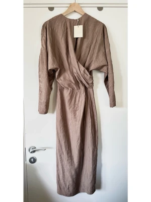 Stylein - helt NY beige / brun klänning med drapering - storlek XXS - Helt NY stilren beige / brun / nougat-färgad klänning från Stylein i storlek XXS. Populära modellen 'Milana' med långa ärmar och en draperad omlottdetalj framtill. Smickrande modell med markerad midja fram och smockdetalj bak för bästa passform. Diskret dold dragkedja baktill och smidig snygg slits nedtill bak för rörlighet. Härligt luftigt finvävt tyg i 88% rayon, så bra naturmaterial som andas. Lika lätt att styla upp till fest som att styla ned till jobb / vardag. En toppenklänning! Helt ny!