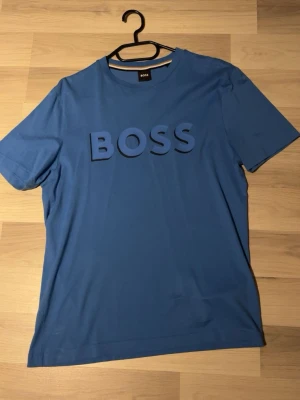 Hugo Boss T-shirt  - Snygg blå t-shirt från BOSS med stort tryck på bröstet där det står 'BOSS'. Klassisk rund halsringning och korta ärmar. Perfekt för en avslappnad och stilren look.