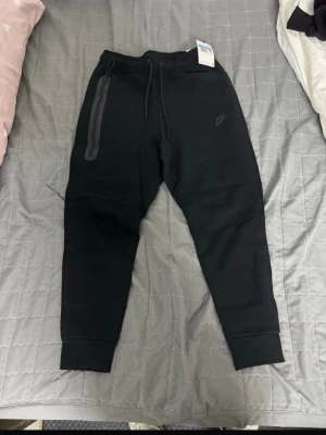 Svarta Nike tech byxor  - Svarta mjukisbyxor från Nike med dragsko i midjan och ribbade muddar vid bensluten. Byxorna har en diskret Nike-logga på låret och en dragkedjeförsedd ficka på ena sidan. Tillverkade i mjukt och bekvämt material, perfekt för en avslappnad stil.