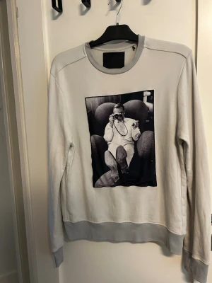 Grå sweatshirt från Limitato - Säljer en vit sweatshirt från Limitato med ett svartvitt fototryck på framsidan. Tröjan har rund hals, ribbade muddar och avslut. Perfekt för dig som gillar unika prints och streetwear-vibe. Finns en liten fläck på ytterkanten av loggan, (se bild 4) men det är inget som syns sålänge man inte kollar noga. Annars mycket fint skick!