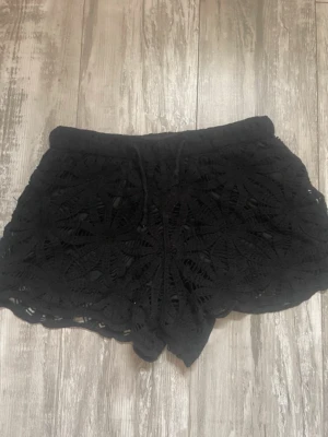 Svarta spetsiga shorts - Snygga svarta shorts med spetsdetaljer. De har en elastisk midja och ett blommigt mönster i spetsen. Perfekta till sommaren och lätta att matcha med olika toppar. Storlek S/M