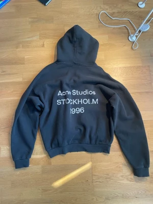 Svart hoodie Acne Studios Stockholm - Svart hoodie från Acne Studios med tryck på ryggen där det står 'Acne Studios STOCKHOLM 1996'. Klassisk modell med huva och ribbade muddar. Perfekt för dig som är 173-180cm.
