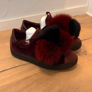 Vinröda Philippe Model sneakers med päls - Unika vinröda sneakers från Philippe Model i mocka med stora fluffiga bollar i röd och svart päls på ovandelen. Skorna har svart sula och detaljer i blankt skinn vid hälen. Perfekt för dig som vill sticka ut med din stil.