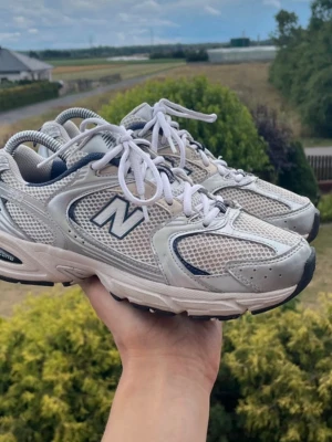 New Balance 530 - Snygga sneakers från New Balance i grått med vita och mörkblå detaljer. Skorna har en sportig design med meshpaneler för extra andningsförmåga och vita snören. Perfekta för en trendig och avslappnad stil.