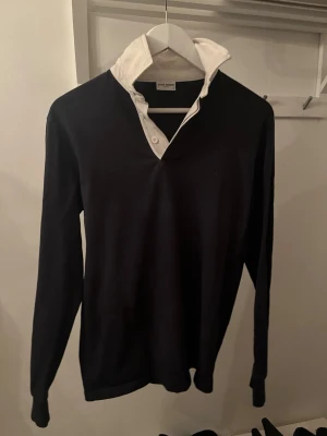 Svart långärmad henley med vit krage från John Henric. - Stilren svart henley med lång ärm och kontrasterande vit krage. Skjortan har knappar vid halsen och en klassisk, enkel design som passar till mycket. Perfekt för dig som gillar en clean look med lite edge.