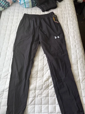 Svarta träningsbyxor från Under Armour - Svarta träningsbyxor från Under Armour med elastisk midja och dragsko. Byxorna har en smal passform och är tillverkade i ett lätt och funktionellt material, perfekt för träning eller sport. Under Armour-loggan syns tydligt på ena benet.