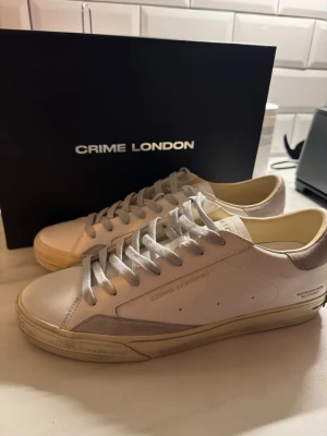 Vita sneakers från Crime London - Stilrena vita sneakers från Crime London med beige detaljer och klassisk låg profil. Skorna har vita snören, gummisula i beige och ovandel i slätt läder. Perfekta för dig som gillar minimalistisk och clean streetstyle.