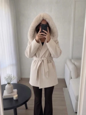 Pälskappa med knytskärp  - Elegant cremefärgad kappa med stor huva och pälsdetaljer. Kappan har ett knytbälte i midjan och praktiska fickor framtill. Perfekt för kyliga dagar när du vill hålla stilen. 