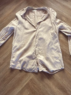 Beige långärmad skjorta - Stilren beige skjorta i mjukt material med klassisk krage och knappar framtill. Skjortan har lång ärm och en avslappnad passform, perfekt att matcha med jeans eller chinos för en enkel look.