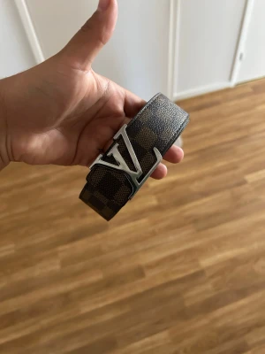 Brunt bälte  - Snyggt bälte i brunt med rutigt mönster Utsidan är i mörkbrunt och ljusbrunt rutigt material, insidan är ljusrosa. Perfekt accessoar för att lyfta din outfit.