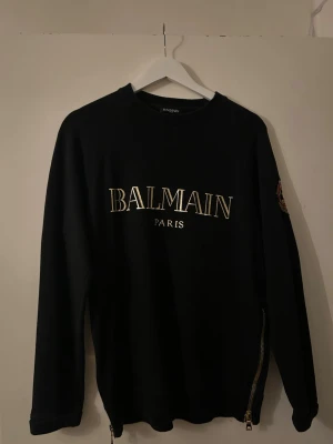 Svart Balmain sweatshirt med guldtryck - Svart sweatshirt från Balmain med stort guldfärgat logotryck på bröstet och broderad patch på ärmen. Tröjan har rund halsringning, långa ärmar och dragkedjedetaljer i nederkant. Perfekt för en trendig och exklusiv streetwear-look.