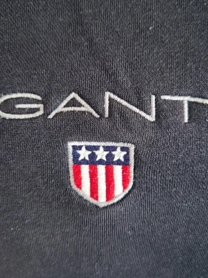 Svart tröja från GANT storlek M - Klassisk svart sweatshirt från GANT med broderad logga och sköld på bröstet. Tröjan har rund halsringning och ribbade muddar vid ärmslut och nederkant. Perfekt för en avslappnad och stilren look.