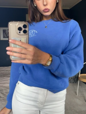Blå sweatshirt med tryck - Snygg blå sweatshirt med vitt tryck på bröstet. Tröjan har rund halsringning, ribbade muddar vid ärmslut och nederkant samt en avslappnad passform. Perfekt till jeans eller kjol för en casual look.