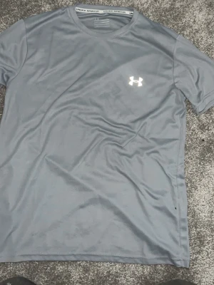 Grå tränings-tshirt från Under Armour - Snygg grå tränings-tshirt från Under Armour med rund hals och korta ärmar. Tillverkad i ett lätt och snabbtorkande material som passar perfekt till gymmet eller löprundan. Diskret logga på bröstet för en sportig look.
