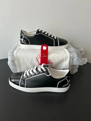 Svarta sneakers från Christian Louboutin - Snygga svarta sneakers från Christian Louboutin med vita kontrastdetaljer och vita skosnören. Skorna har en klassisk låg silhuett och den ikoniska röda sulan. Tillverkade i blankt läder med logga på hälen och sidan. Perfekta för dig som vill sticka ut.  Endast OG dustbag och skohorn medföljer.