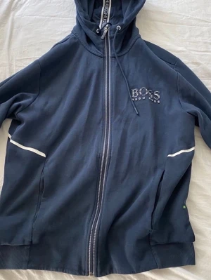 Marinblå hoodie från Hugo Boss - Snygg marinblå hoodie från Hugo Boss med dragkedja framtill och vit logga på bröstet. Hoodien har huva med dragsko och vita detaljer längs ärmarna. Perfekt för en avslappnad och stilren look.