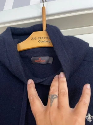 Mörkblå stickad hoodie från Morris - Snygg mörkblå stickad hoodie från Morris med dragsko i huvan och diskret logga på bröstet. Tröjan har långa ärmar och ribbade muddar vid ärmslut och nederkant. Mamma har tvättat den så den sitter mer som en S men inga defekter finns på tröjan 