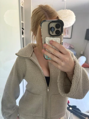 Beige teddyjacka med dragkedja - Beige teddyjacka med dragkedja framtill och två stora fickor. Jackan har en hög krage och är gjord i fluffigt, mjukt material som ger en avslappnad och trendig look. Perfekt för kyliga dagar och enkel att matcha med olika outfits.