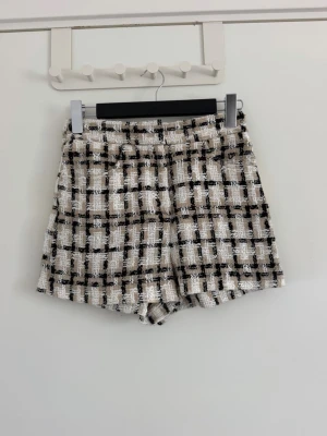 Rutig tweedshorts i beige och svart - Snygga shorts i tweed med rutigt mönster i beige, svart och vitt. De har en högmidjad passform och en lite strukturerad yta som ger en exklusiv känsla. Perfekta för dig som vill sticka ut med en trendig och chic stil. Använda en gång, köpta från Sellpy 