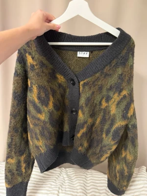 Leopardmönstrad kofta från STAY - Säljer en fluffig kofta från STAY med leopardmönster i grönt, gult och svart. Koftan har bred ribbstickad kant vid hals, ärmslut och nederkant samt knäppning med svarta knappar framtill. Oversized passform och mysigt material.