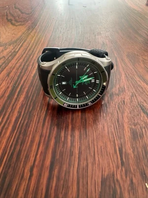 Svart dykarklocka med grön urtavla - Snygg dykarklocka med svart urtavla och gröna detaljer på visarna och ringen. Boetten är i silverfärgad metall och armbandet är svart, troligtvis i syntetmaterial. Klockan har datumvisning och tydliga markeringar runt urtavlan. Perfekt för dig som gillar stilrena accessoarer.