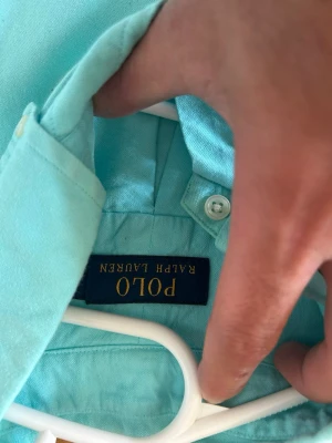 Turkos skjorta från Polo Ralph Lauren - Snygg turkos skjorta från Polo Ralph Lauren med klassisk krage och knäppning framtill. Skjortan har den ikoniska röda loggan broderad på bröstet och är tillverkad i ett lätt och bekvämt material, perfekt för en fräsch och stilren look.