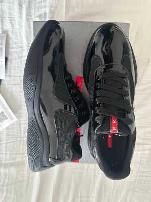 Svarta sneakers från Prada - Snygga svarta sneakers från Prada med glansig finish och meshdetaljer. Skorna har svarta skosnören och en röd logotyp på plösen. Sulan är svart och ger ett sportigt intryck. Perfekta för dig som vill ha en stilren och exklusiv look.