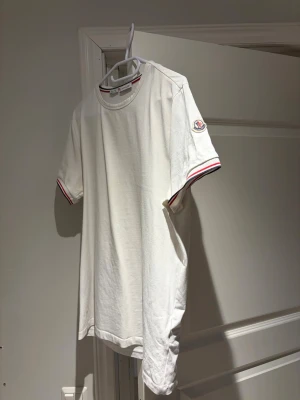 Vit Moncler t-shirt med rödblå detaljer - Vit t-shirt från Moncler med korta ärmar och rund hals. Ärmsluten och kragen har snygga rödblå ränder och Moncler-logga sitter på vänster ärm. Tillverkad i mjuk bomull för en skön känsla och clean look.