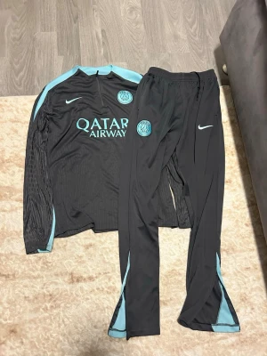 Svart PSG träningsset Nike - Säljer ett svart träningsset från PSG och Nike med ljusblå detaljer. Setet består av långärmad tröja och byxor, båda med PSG-logga och Nike swoosh. Tröjan har Qatar Airways-tryck på bröstet och dragkedja vid halsen. Perfekt för fotbollsträning.