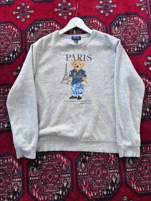 Grå sweatshirt Polo Ralph Lauren - Grå sweatshirt från Polo Ralph Lauren med ikoniska björnen på bröstet, texten 'PARIS' och Eiffeltornet i bakgrunden. Tröjan har rund halsringning och ribbade muddar. Perfekt för dig som gillar streetwear och klassiska prints. Finns ej tags inne i tröjan. Storlek: XL 18-20, dvs. herr M. 