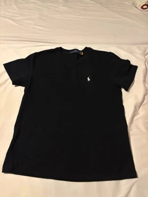 Svart t-shirt från Polo Ralph Lauren - Klassisk svart t-shirt från Polo Ralph Lauren med rund hals och korta ärmar. Diskret broderad logga i vitt på bröstet. Tillverkad i mjuk bomull för en skön och avslappnad stil.